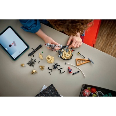 Peças LEGO coloridas e tablet com instruções, criança montando brinquedo de teia de aranha numa mesa cinza