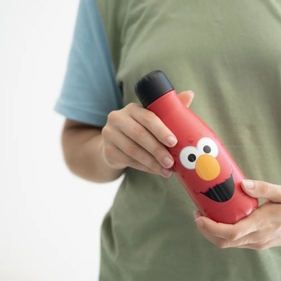 Garrafa térmica vermelha com rosto do Elmo e tampa preta nas mãos de uma pessoa com camiseta azul e verde.