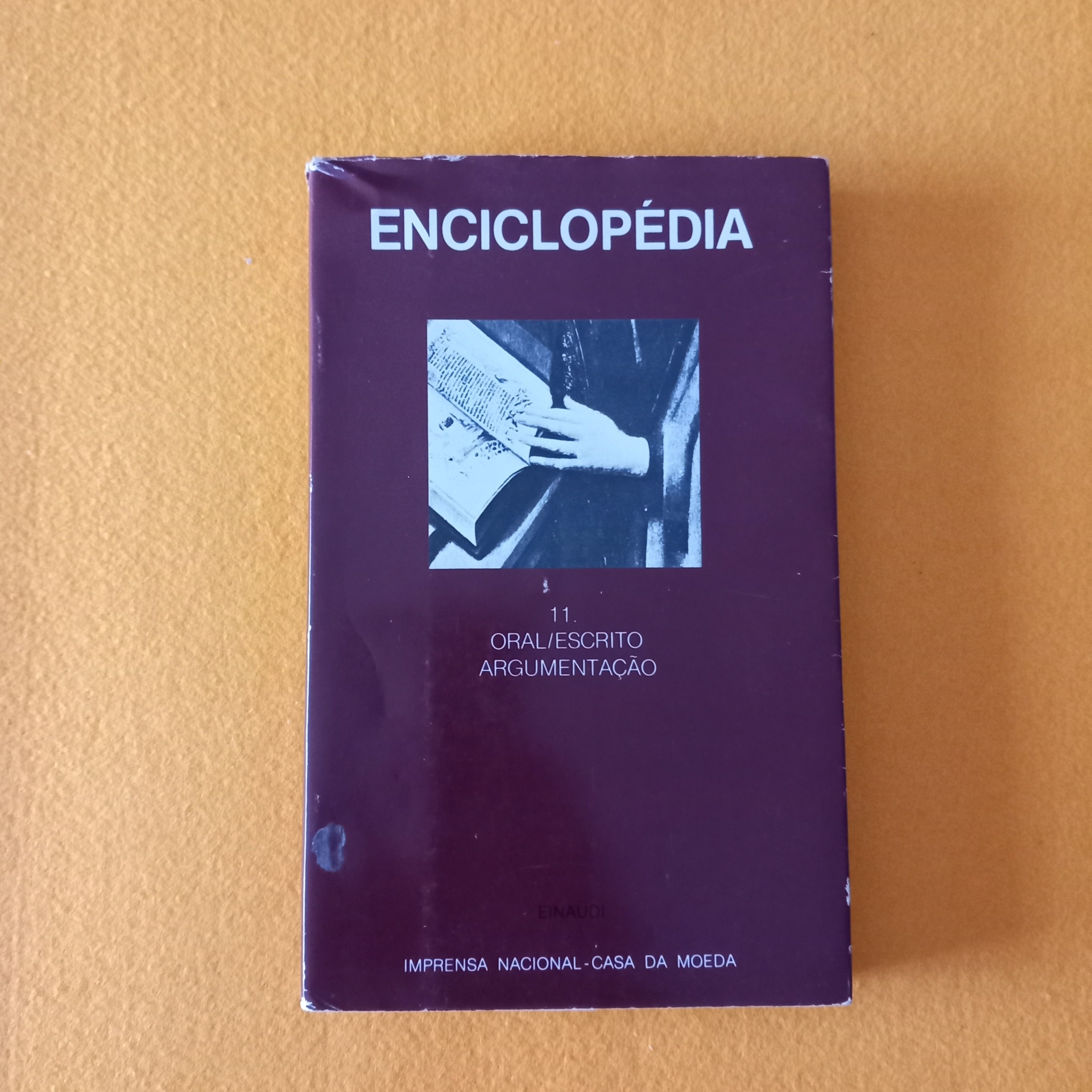 Enciclopédia Einaudi: 11 Oral/Escrito, Argumentação Livro ENCICLOPÉDIA com capa roxa e imagem preto e branco sobre superfície laranja