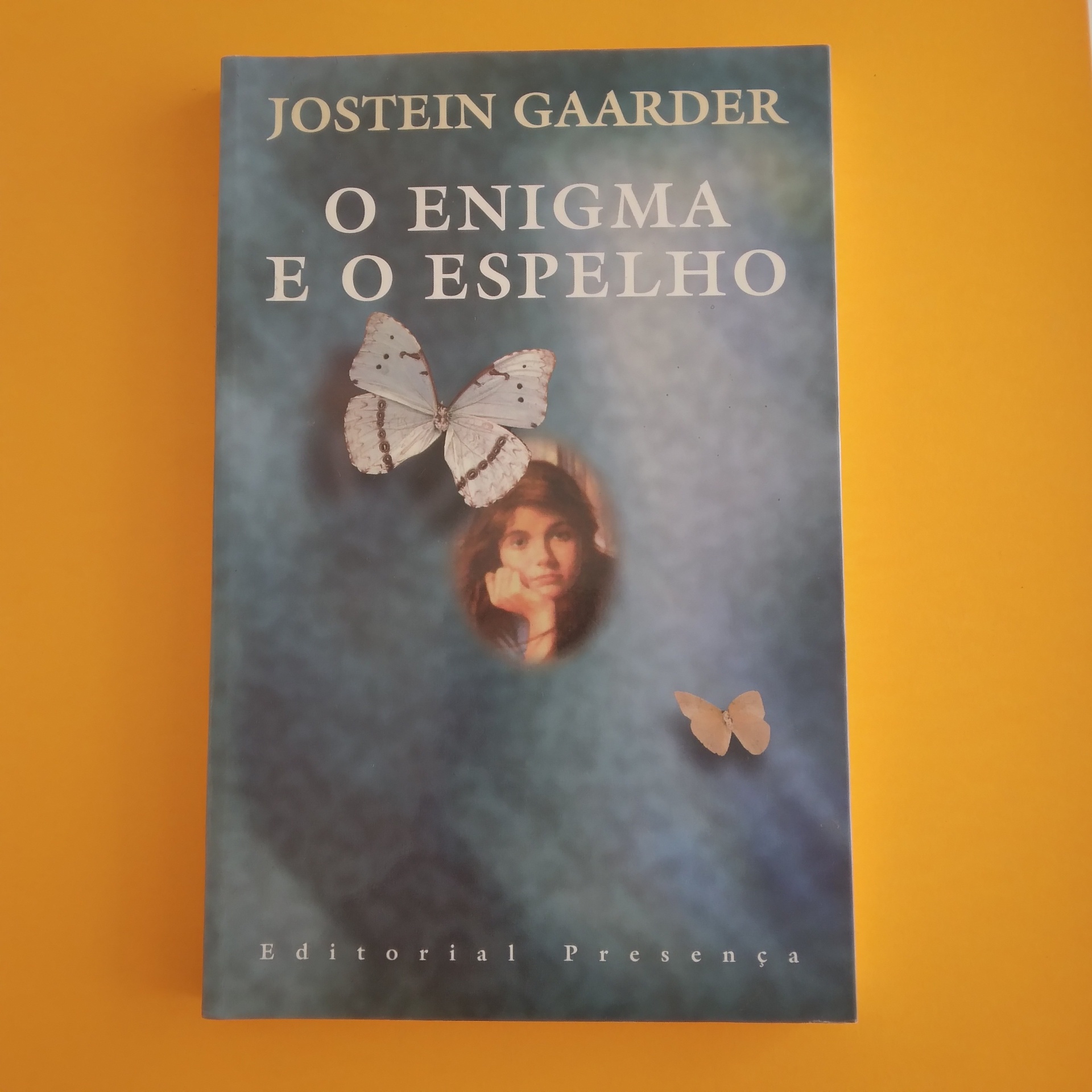 O Enigma do Espelho, Jostein Gaarder Capa de livro azul com borboletas e rosto de criança, texto 'O ENIGMA E O ESPELHO', autor JOSTEIN GAARDER