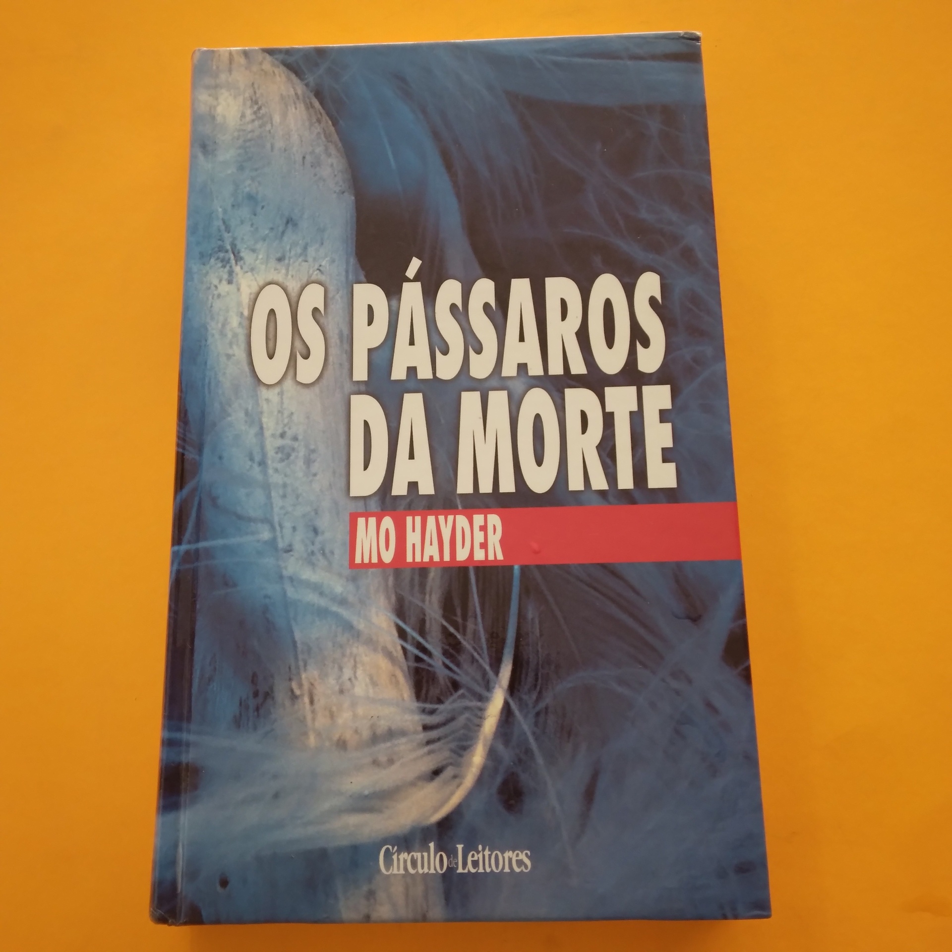 Os Pássaros da Morte, Mo Hayder Livro 'Os Pássaros da Morte' de Mo Hayder com capa azul e branca e penas sobre fundo amarelo