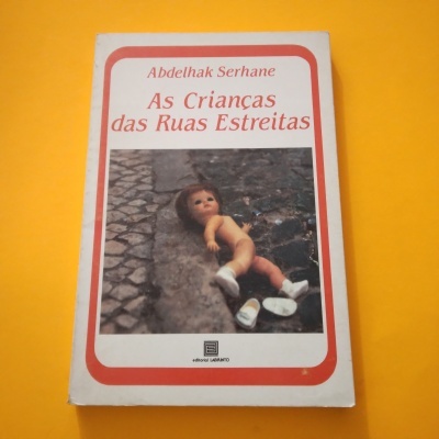 Capa de livro com fotografia de boneca numa rua de calçada sobre fundo amarelo