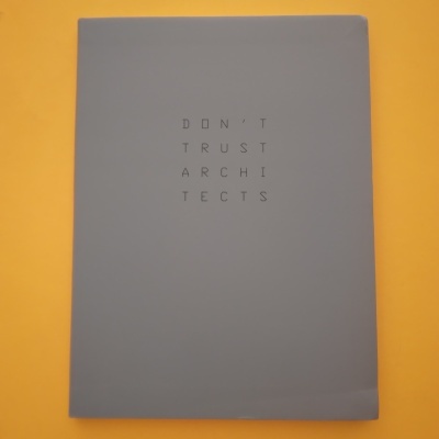 Livro cinzento com texto DON'T TRUST ARCHITECTS numa superfície laranja