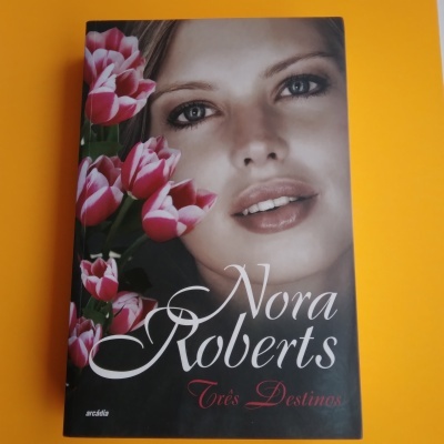Capa de livro com rosto de mulher e flores cor-de-rosa, fundo escuro, texto Nora Roberts Três Destinos