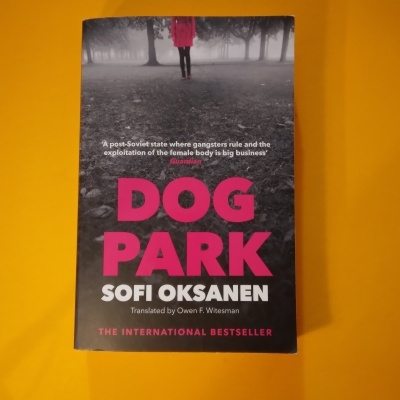 Capa do livro Dog Park de Sofi Oksanen com texto em rosa e branco em fundo de parque enevoado.