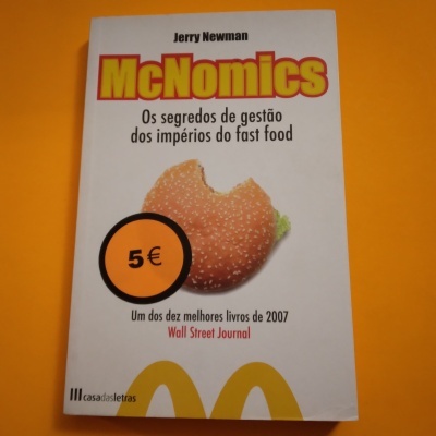 Livro McNomics com capa branca, pão de hambúrguer mordido, preço 5€ e fundo amarelo.