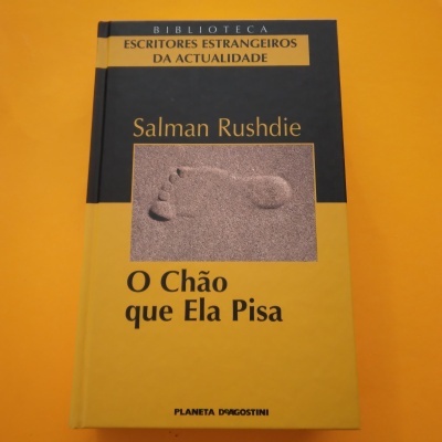 Capa de livro 'O Chão que Ela Pisa' de Salman Rushdie com cores amarela e preta e imagem de pegada na areia