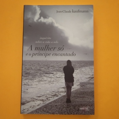 Capa de livro com mulher de costas à beira-mar em preto e branco sobre fundo laranja