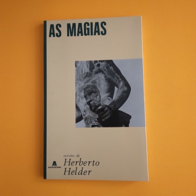 Livro 'AS MAGIAS' com capa cinza e imagem de escultura, sobre fundo laranja