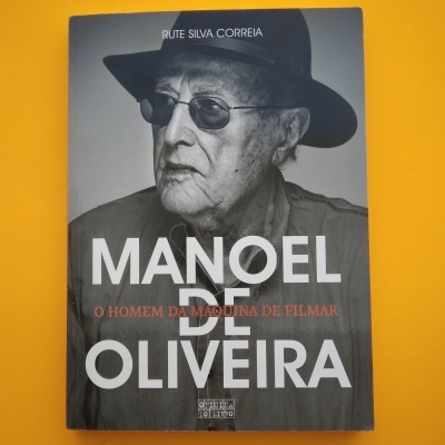 Capa de livro com fotografia em preto e branco de homem idoso com chapéu e óculos escuros, título MANOEL DE OLIVEIRA em branco, subtítulo em laranja e autor RUTE SILVA CORREIA.