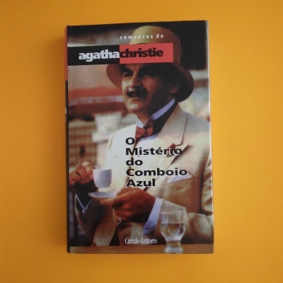 Livro 'O Mistério do Comboio Azul' de Agatha Christie com capa com homem de fato bege e chapéu castanho segurando chávena