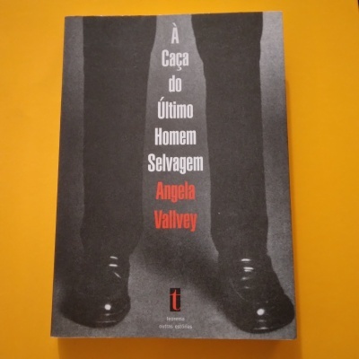 Capa do livro 'À Caça do Último Homem Selvagem' em preto e branco com texto e imagem de pernas e sapatos pretos