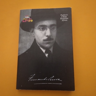 Capa de livro preto com foto em preto e branco de Fernando Pessoa usando chapéu e óculos