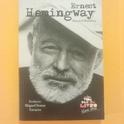 Capa do livro Ernest Hemingway com fotografia preto e branco de um homem idoso de barba e chapéu em fundo amarelo.