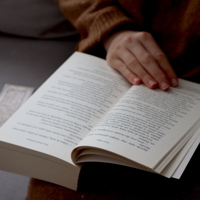 Pessoa segurando um livro aberto com texto em português, sentada num ambiente interior.