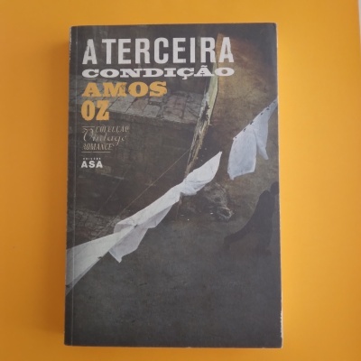 Capa do livro A Terceira Condição de Amos Oz com fundo cinza e texto em branco e amarelo.