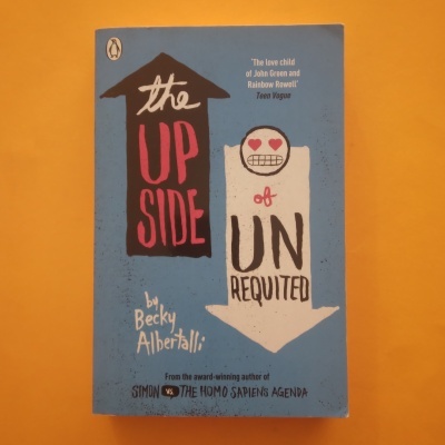 Capa do livro azul 'The Upside of Unrequited' com setas e desenho de rosto em fundo laranja