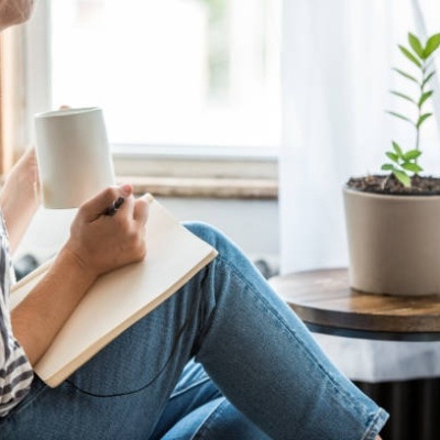 Mulher sentada com caneca branca e caderno junto a janela com planta em vaso