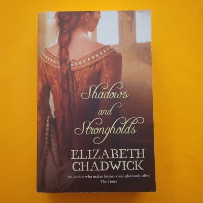 Capa de livro 'Shadows and Strongholds' com imagem de mulher vestida à época em fundo amarelo.