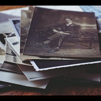 Pilhas de fotografias antigas a preto e branco sobre uma mesa de madeira com papéis manuscritos ao fundo