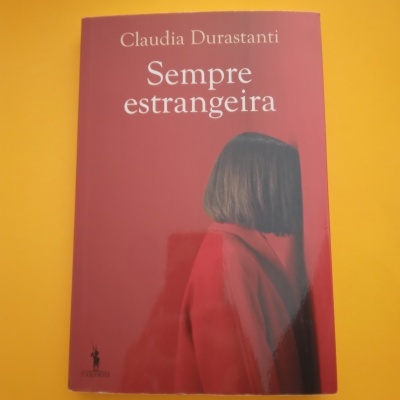 Capa de livro 'Sempre estrangeira' de Claudia Durastanti, pessoa com cabelo curto e casaco vermelho sobre fundo amarelo.