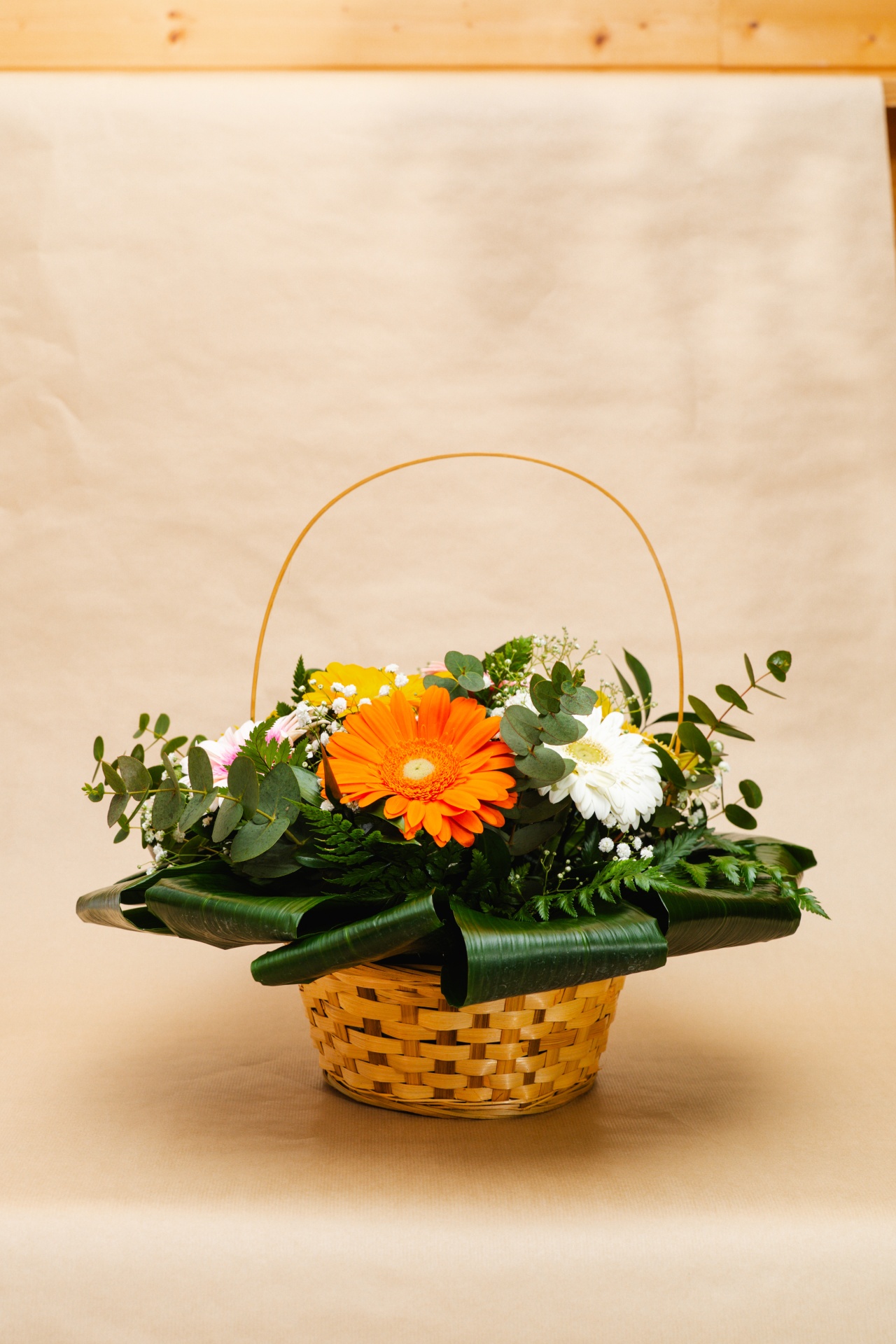 Cesta de flores com gerberas laranja, branca e amarela e folhas verdes em fundo bege.