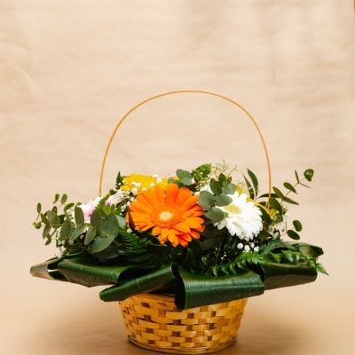 Cesta de flores com gerberas laranja, branca e amarela e folhas verdes em fundo bege.