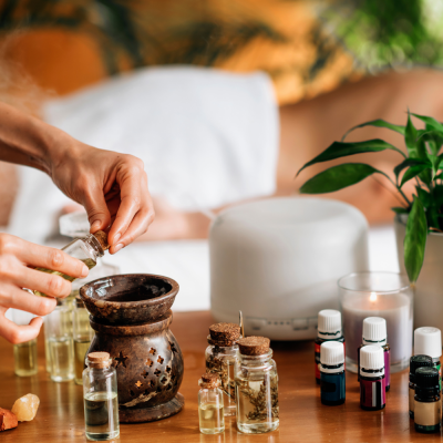 Frascos de óleos essenciais, queimador cerâmico castanho, vela e difusor branco numa mesa com planta verde