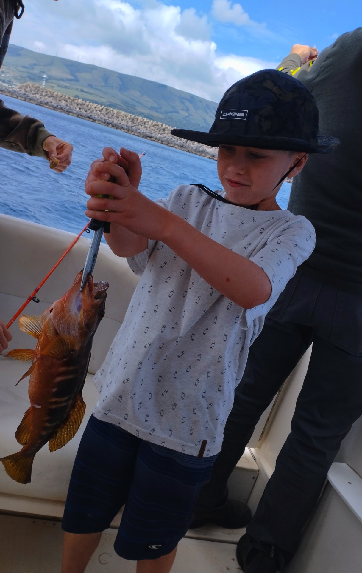 Criança com chapéu preto segurando peixe num barco com mar e montanhas ao fundo