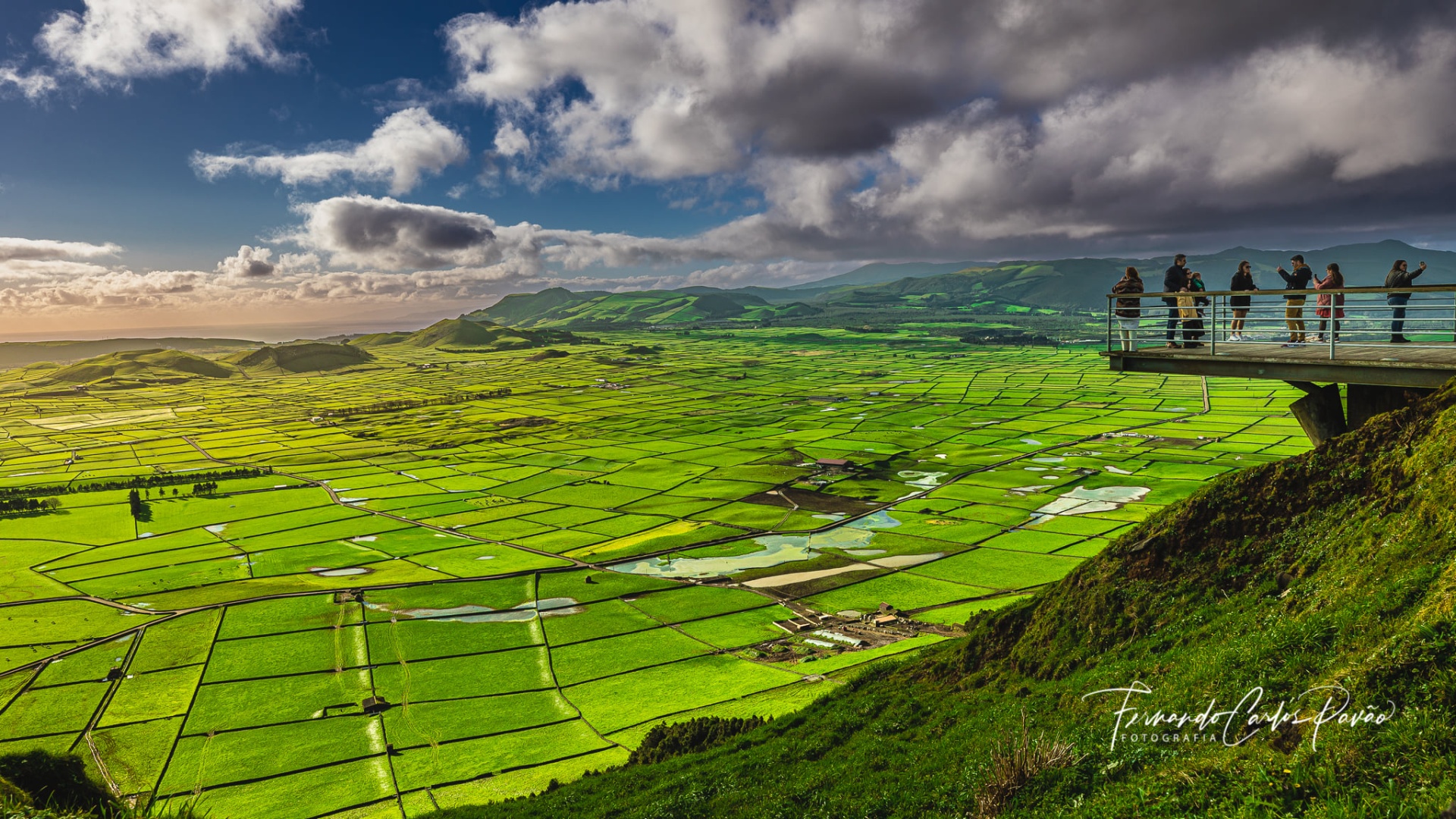 Campos verdes agrícolas vistos de uma colina com pessoas em plataforma de observação sob céu nublado