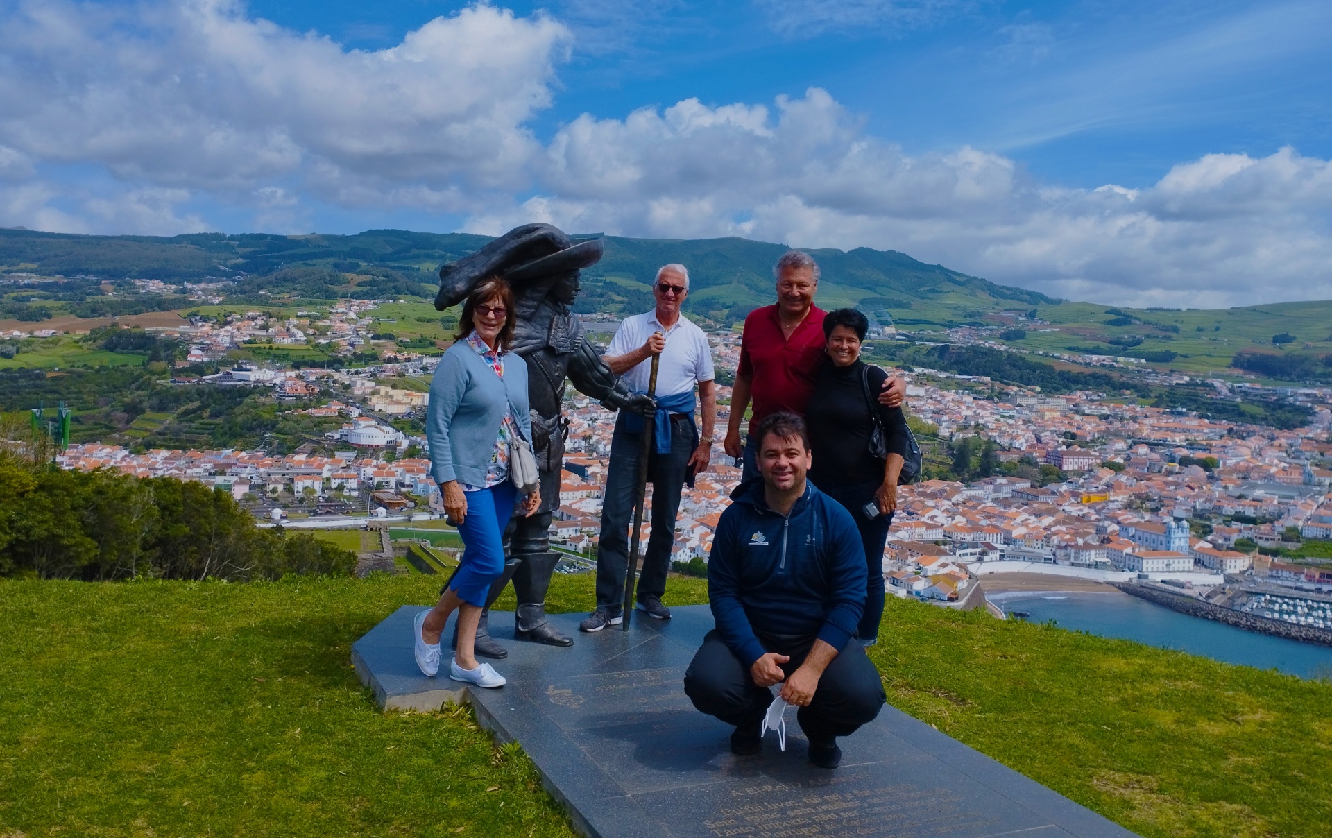 Cinco pessoas junto a estátua de bronze numa área verde com vista para cidade e mar