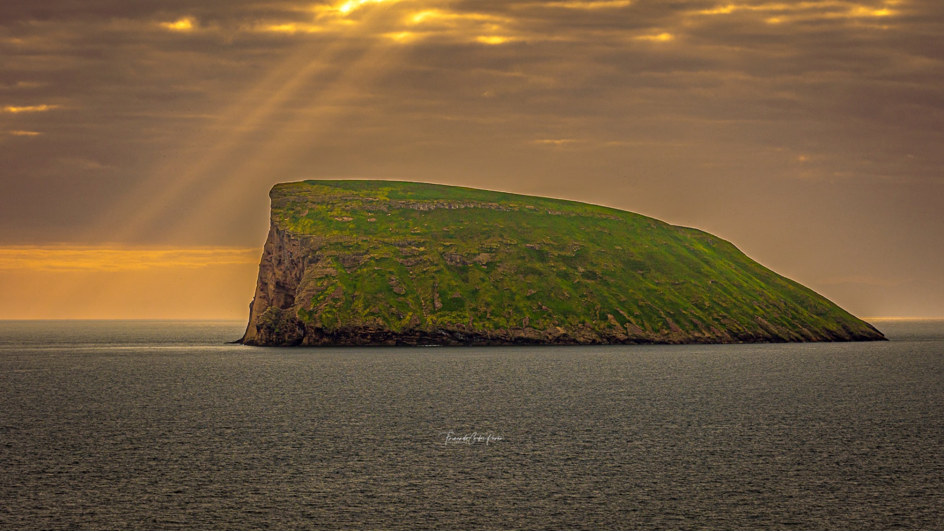 Ilha rochosa verde rodeada de água sob céu com raios de sol