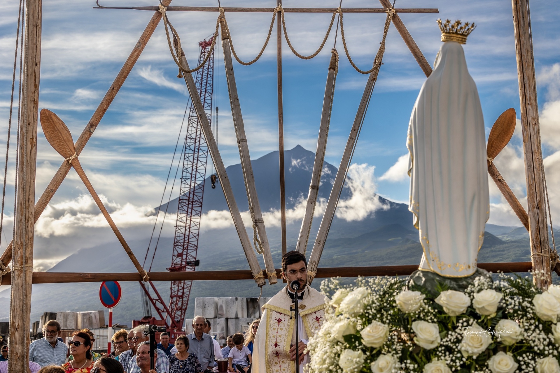 Procissão religiosa com padre e estátua da Virgem Maria coroada junto a flores brancas ao ar livre