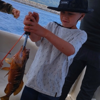 Criança com chapéu preto segurando peixe num barco com mar e montanhas ao fundo