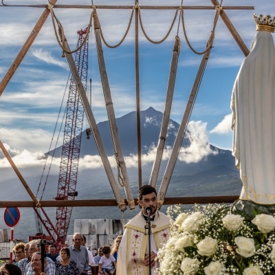 Procissão religiosa com padre e estátua da Virgem Maria coroada junto a flores brancas ao ar livre
