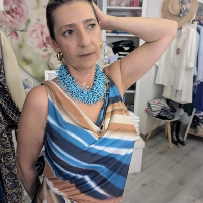 Mulher com vestido listrado azul e bege e colar azul numa loja de roupas decorada com flores e chapéus