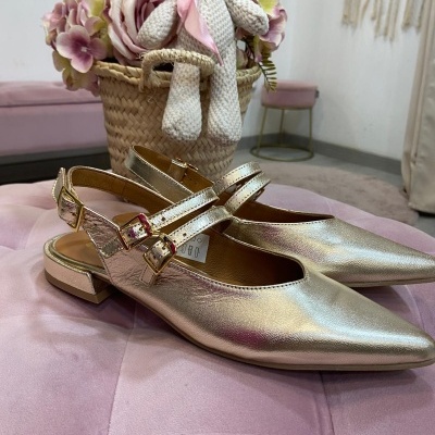 Sapatos dourados com tiras e fivelas sobre puff rosa com cesto e urso pelúcia ao fundo