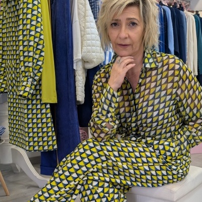 Mulher com fato geométrico amarelo, branco e preto sentada em cadeira branca entre roupas penduradas