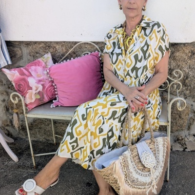 Mulher com vestido estampado sentada num banco branco com almofadas cor-de-rosa, sandálias brancas e mala de palha com franjas.