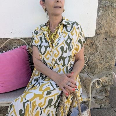 Mulher com vestido comprido estampado em verde, amarelo e branco, sentada num banco de ferro junto a almofada cor-de-rosa