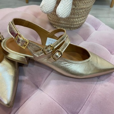 Sapatos femininos dourados com fivela e salto baixo sobre almofada rosa.