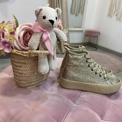 Ursinho de crochê em cesto de vime com flores e ténis dourados em superfície almofadada cor-de-rosa