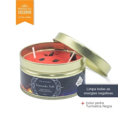Vela aromática vermelha em lata dourada com tampa ao lado, rótulo negro com texto e pedras pretas na vela.