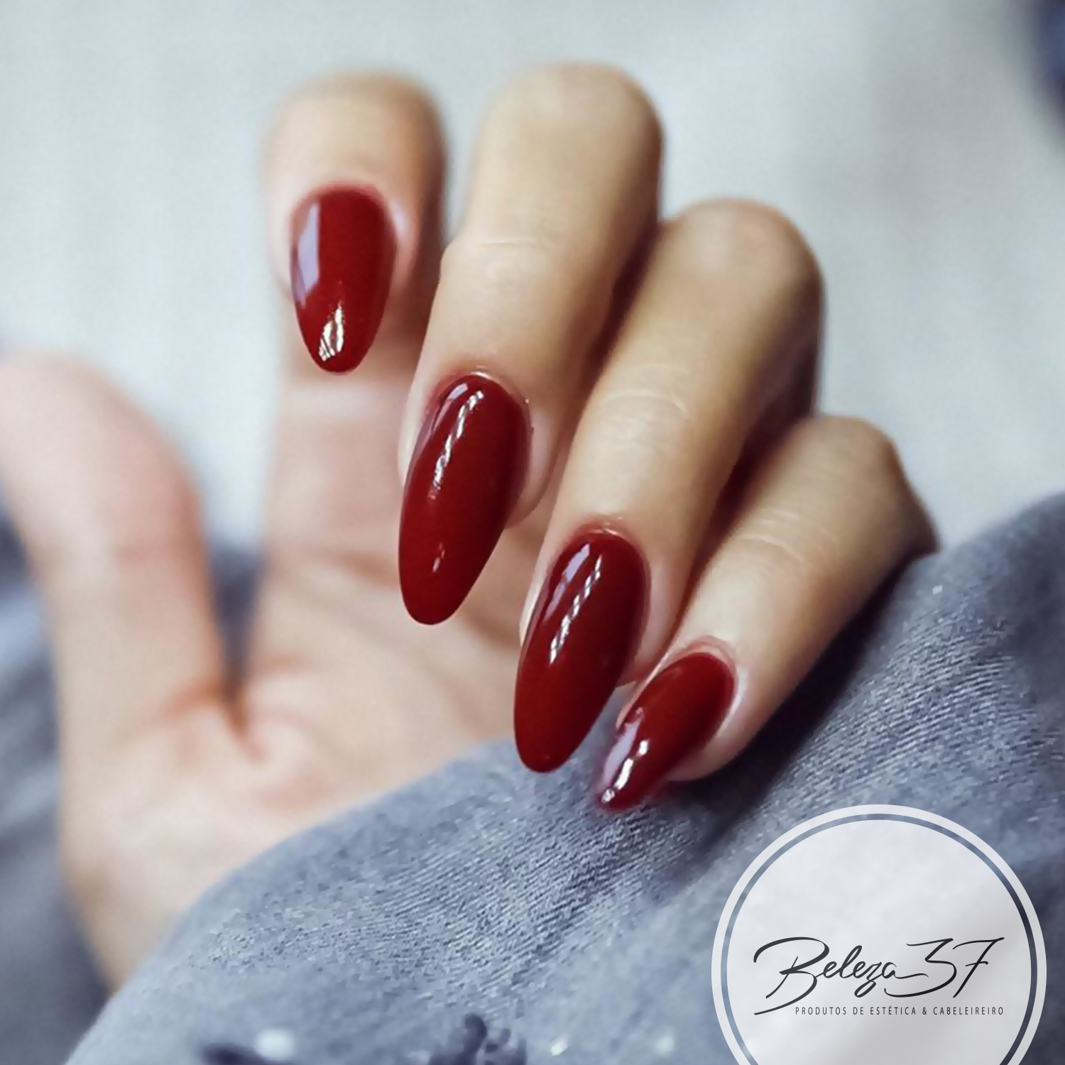 Verniz Gel Andreia 297 – O vinho (bordeaux) Unha artificial longa pintada com verniz vermelho brilhante em fundo desfocado.