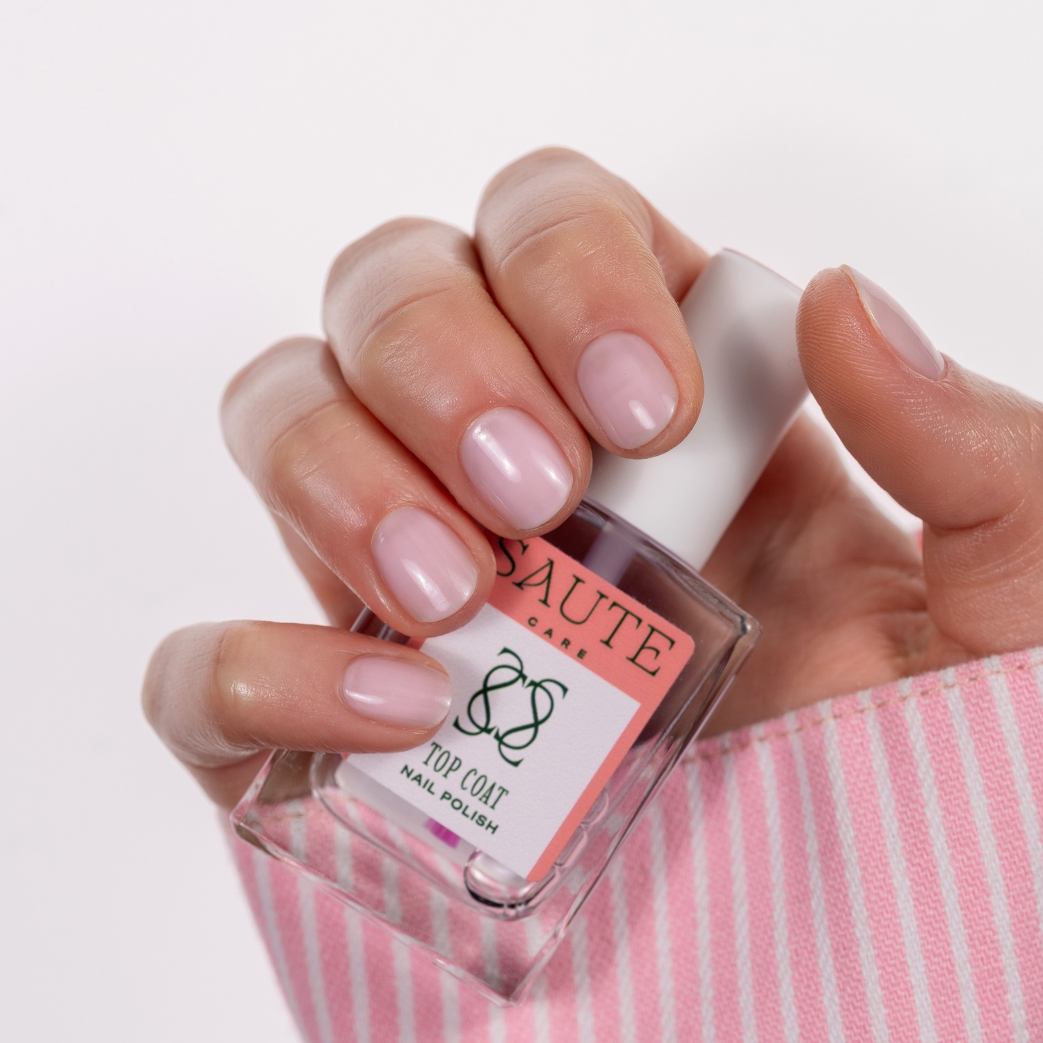 Top Coat Saute Care 15 ml – Proteção e Brilho para Manicure Tradicional Frasco de verniz de unhas Saute Care Top Coat segurado por mão com unhas rosa claro.