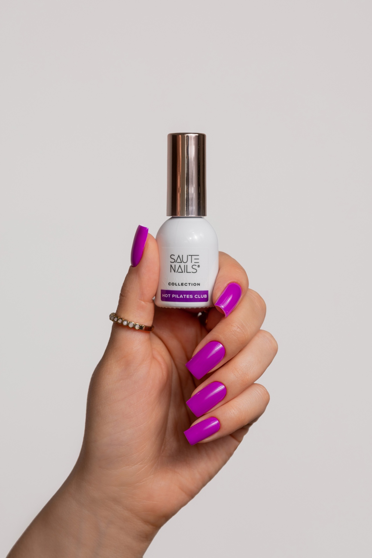 Verniz Gel Hot Pilates Club (Fúcsia‑Violeta Néon) Mão segurando frasco de verniz de unhas branco com tampa metálica e texto da marca.