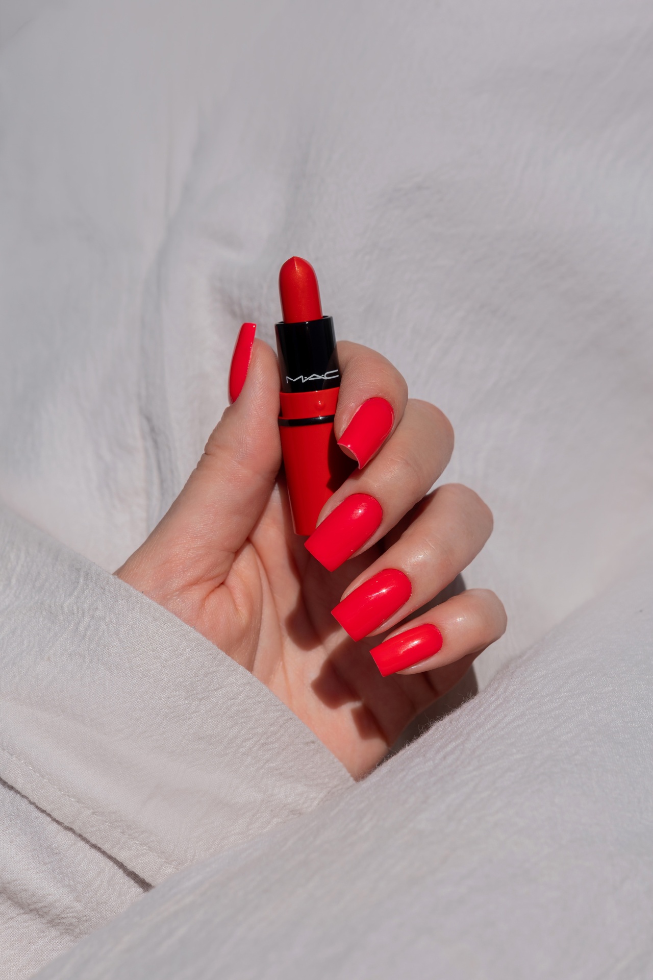 Verniz Gel Red Light Baby (Vermelho Laranja Néon) Batom vermelho MAC segurado por mão com unhas vermelhas brilhantes em fundo branco