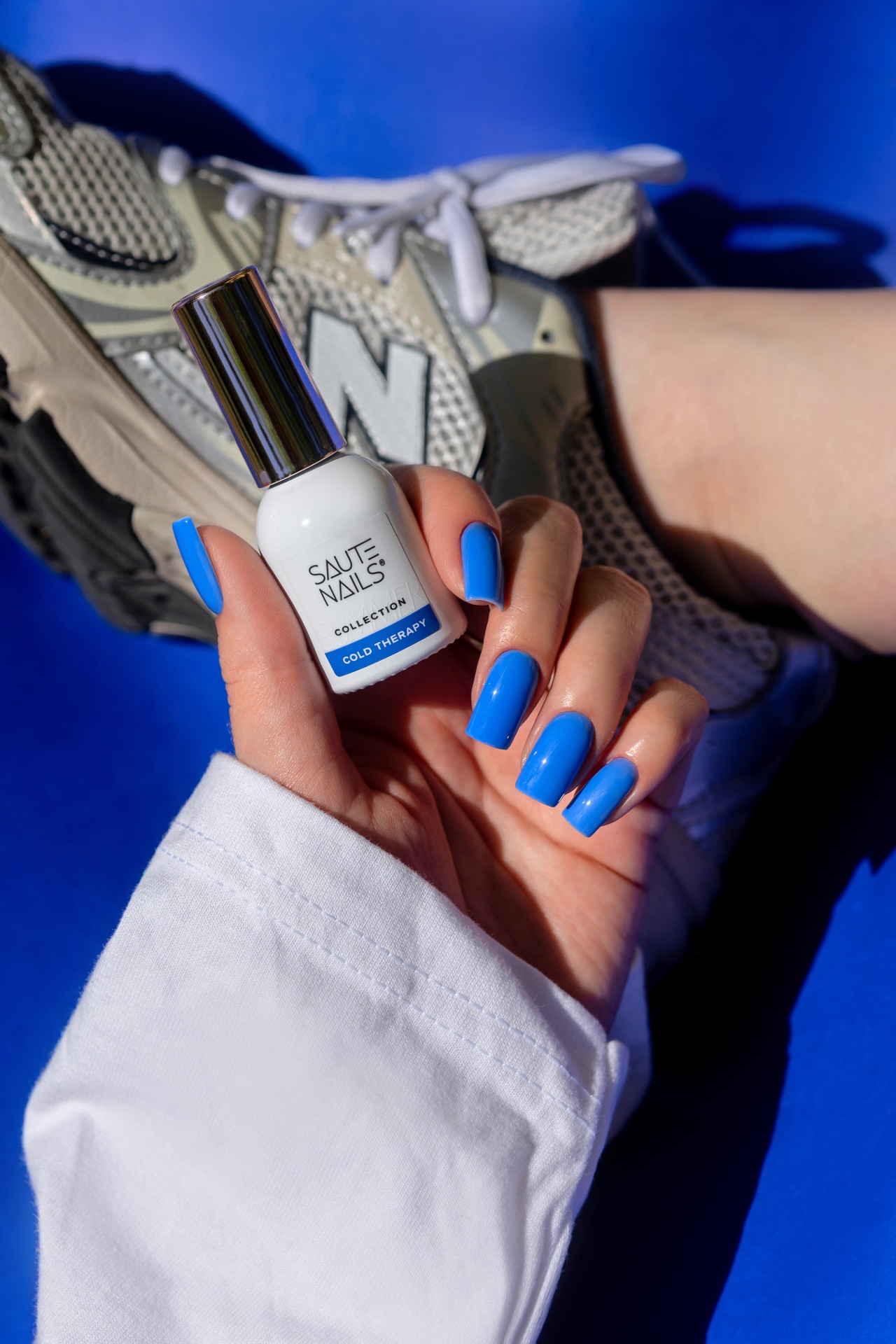 Verniz Gel Cold Therapy (Azul Royal Intenso) Frasco branco de verniz Saute Nails Collection Cold Therapy com unhas azuis e sapato desportivo cinzento ao fundo