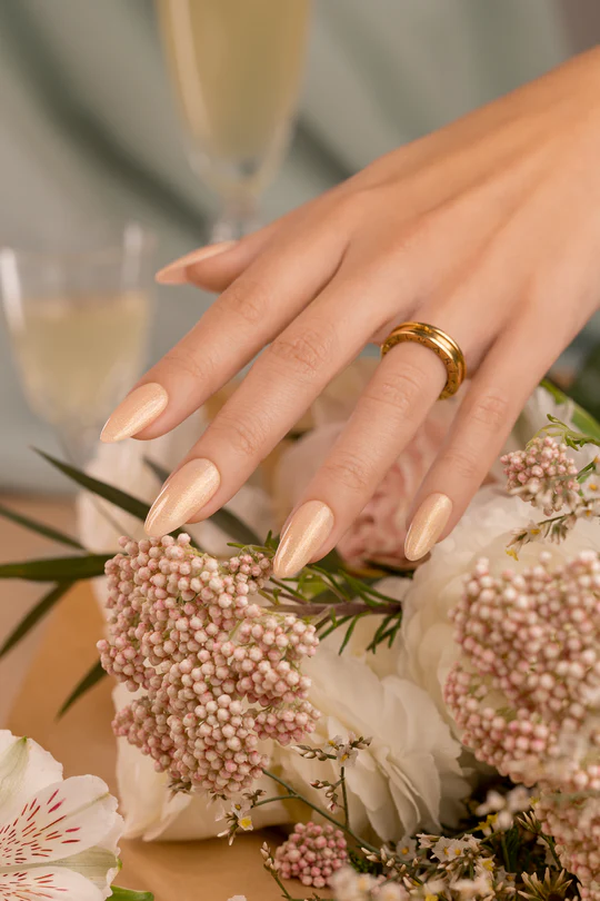 Fusion Color ID3 - Wedding Toast - Nude champagne com brilhos dourado Mão feminina com unhas bege creme usando anel dourado junto a flores brancas e cor de rosa