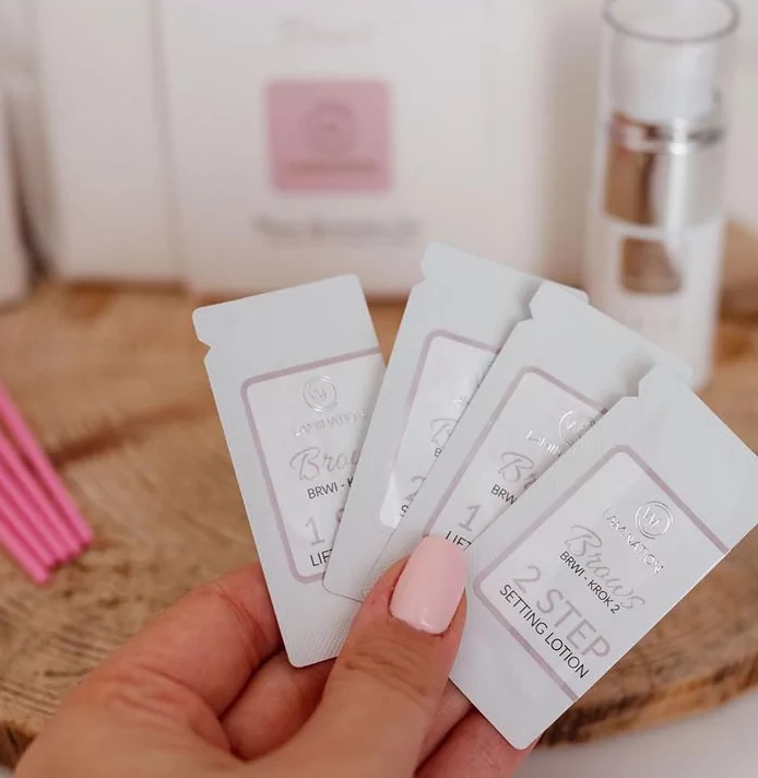 Lifting Sobrancelhas Wonders Lashes - Passo 1 Mão segurando amostras brancas de cosméticos Brown Kross com texto visível, fundo com frascos e caixa em madeira